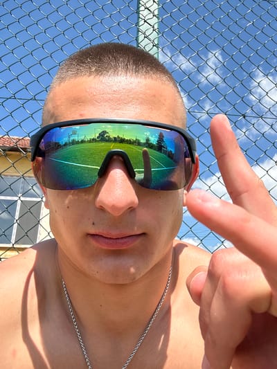 A close-up, rotated image depicts a light-skinned individual with a shaved head wearing sunglasses reflecting a sports field with a basketball hoop. The person's left hand with two fingers extended is visible in the upper portion of the frame. They are standing near a chain-link fence against a partly cloudy blue sky. The lower portion of the frame shows a building with a visible window and brown tiled roof, alluding to the possible location of Kalchevo, Bulgaria. The person wears a silver necklace. The lighting suggests a sunny day.