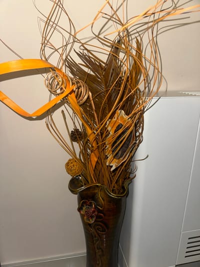 The image displays an indoor scene in Radishevo, Bulgaria, featuring a tall, dark brown, ornate floor vase. The vase has a wavy upper rim and an embossed reddish-brown floral design on its upper body, with additional textured patterns extending vertically. The vase contains a large decorative arrangement of dried botanical elements. This arrangement includes numerous long, thin, curved brown and orange twigs, wider dark brown palm fronds, and flat strips of orange material formed into spirals and angular shapes. Additional components visible within the arrangement are small, woven light brown spheres, pinecones, and dried, light-colored shell-like or seed pod elements with dark centers. The vase stands against a plain, light-colored wall on the left, and a white rectangular unit with horizontal vents, possibly an air conditioning unit or radiator cover, on the right. A narrow section of gray flooring is visible at the bottom of the frame. No individuals are present in the scene.