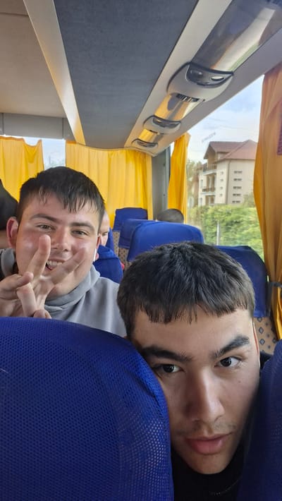 This image captures two young men seated inside a bus, likely in Krivodol, Bulgaria, as indicated by the presence of a distinctive residential building visible through the window. The foreground is dominated by the heads and shoulders of the two individuals, with the person on the left looking directly at the camera and making a peace sign with their right hand. They are wearing a grey hoodie. The person on the right is closer to the camera and their face is partially obscured by a blue seat. The interior of the bus features blue seats and yellow curtains, with overhead compartments visible. Outside the window, a multi-story building with a beige facade and dark windows is seen, surrounded by greenery. The sky appears overcast.