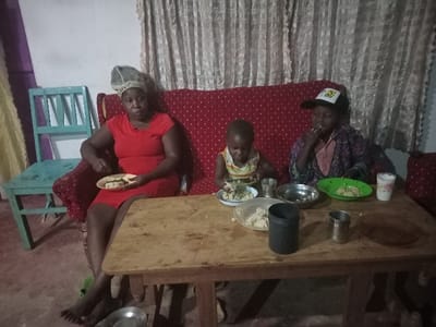 Three individuals sit around a wooden table, seemingly enjoying a meal. A woman in a red dress sits to the left, holding a plate with food. To her right, a young child in a striped tank top eats from a bowl. Next to the child, another person wearing a baseball cap is also eating. Several plates and cups are scattered across the table. A red patterned sofa serves as a backdrop, with a curtain visible behind it. A blue wooden chair is on the left side of the image.