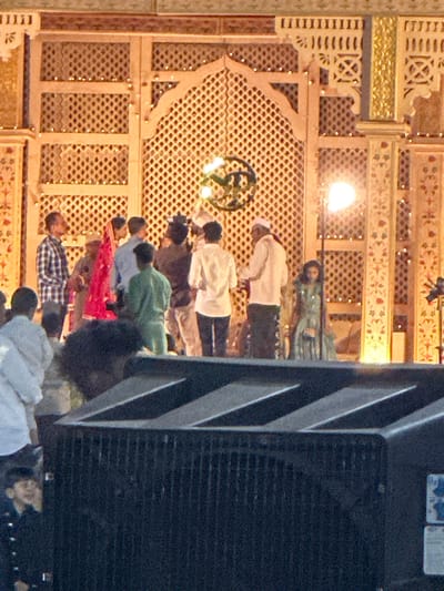 A group of people are praying on the floor inside the Hazrath Sayyed Khwaja Meer Sayyed Ali Dargah, located in Nashik, India. The prayer area has a beige latticework and ornate gold details. Some of the people are wearing white robes, others are wearing various colored shirts, including green, red, and plaid. An air conditioning unit can be seen on the right side of the image.