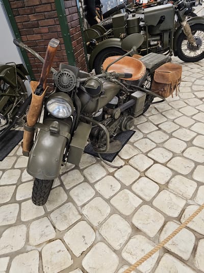 A high-angle shot shows vintage military motorcycles on display in Архангельское, Russia. The motorcycles are painted in drab olive green, with brown leather accents like saddlebags and handlebar covers. The motorcycle in the foreground is the most prominent, revealing detailed engine components, a headlight, and a front fender. Behind, another motorcycle is partially visible, stacked higher than the first. The display area features a faux brick floor with a green-painted border. The backdrop is a wall made of large, irregularly shaped stones in a light beige color. A thick rope, possibly part of a barrier, is visible in the upper right.