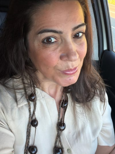 A woman with dark brown hair is facing the camera while inside a car. She is wearing a light-colored, collared shirt and a two-strand necklace made of dark beads. The top of the car window is visible in the upper left corner. No specific details indicate the location is Niederanven, Luxembourg.