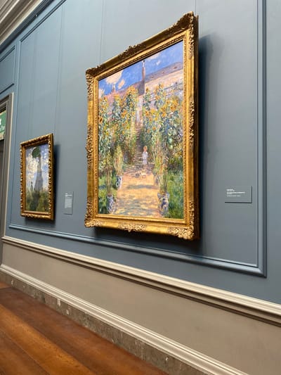 An indoor museum setting shows two Impressionist paintings displayed on a muted blue wall. The painting on the right, larger and closer to the viewer, depicts a sun-drenched garden with a pathway lined with tall sunflowers and other flowers. A small child and a dog are visible on the path. The painting is framed in an ornate gold frame. To the left and slightly behind is a smaller painting of a woman with an umbrella standing in a garden. Both paintings are hung on a wall with decorative molding near the ceiling and a lighter colored wainscoting below. A plaque with text is visible to the right of the larger painting. The floor is made of polished wooden planks. The location is the National Gallery of Art in Washington, D.C., United States of America.