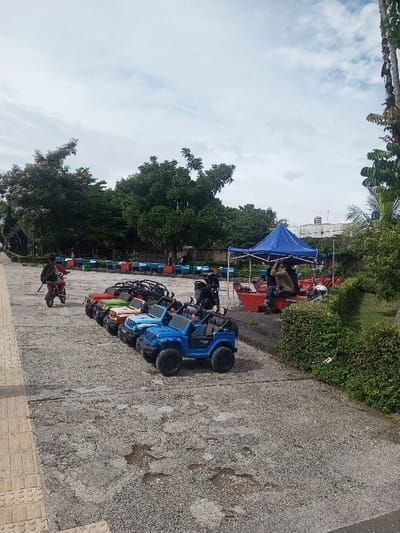 A row of toy jeep-like vehicles in bright colors sit on a cracked pavement. A blue tent with people underneath stands nearby, alongside a row of colorful benches. A child rides a scooter in the background. Lush green trees and bushes surround the scene, and the sky is partly cloudy. The vehicles are the main focus, with the environment providing context.