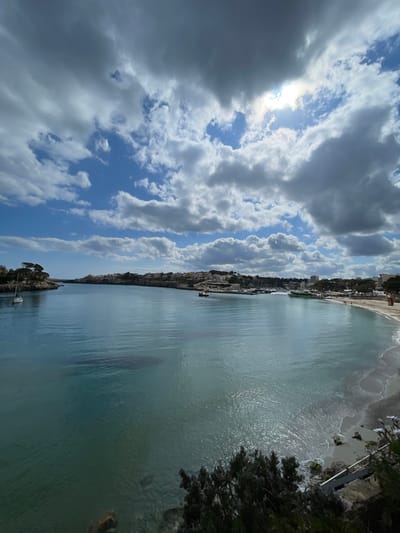This image depicts the Bay of Porto Cristo, Spain, viewed from an elevated position. The sky is partially covered by cumulus clouds, with significant patches of bright blue visible, particularly towards the left. The sun is obscured by a dense cloud formation in the upper right quadrant, casting a bright glare through a clearing.
The bay's water is a translucent turquoise and blue, appearing calm with minimal surface ripples. In the mid-ground, a white sailboat with a single mast is anchored near the left entrance of the bay, a rocky, tree-covered headland. Further into the bay, towards the right, a marina or port area is visible, containing multiple moored vessels, including a prominent green and white catamaran-style boat.
The right side of the bay features a crescent-shaped sandy beach, leading to an urbanized shoreline. Several indistinct figures, presumed to be individuals, are present on the beach. Buildings, varying in height and density, line the coast, indicating the town of Porto Cristo.
In the immediate foreground, at the bottom right, dark green foliage from bushes or trees partially extends into the frame. No specific actions or interactions are clearly discernible beyond the general presence of boats and individuals on the beach.