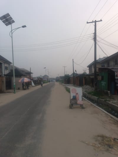 A street view showcases a somewhat desolate scene. The road, displaying signs of wear, stretches into the distance under a hazy sky. Buildings line either side, a mix of residential structures and small businesses. Utility poles with a web of wires add to the unkempt aesthetic. A solar-powered street lamp stands tall on the left. A few individuals are visible; one under an umbrella stand and another is riding a bicycle. There's also garbage on the side of the road.