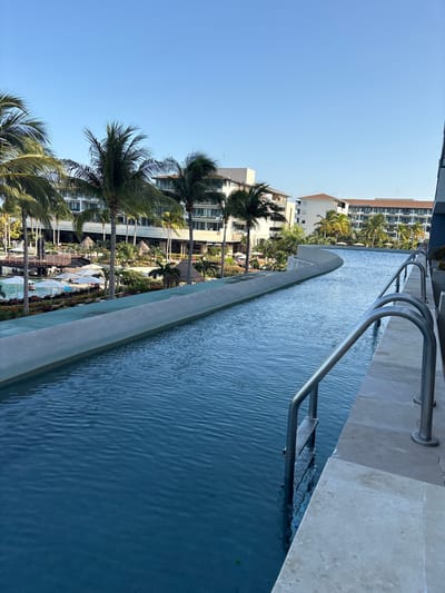 The image depicts a resort swimming pool area in Punta Sam, Mexico. A long, winding swimming pool with clear blue water occupies the foreground and middle ground. Stainless steel railings and ladders are visible along the edge of the pool. Beyond the pool, palm trees line the area, with a resort building visible in the background. There are lounge chairs and umbrellas near a section of the pool, and a few people are seen relaxing. The sky is clear and blue.