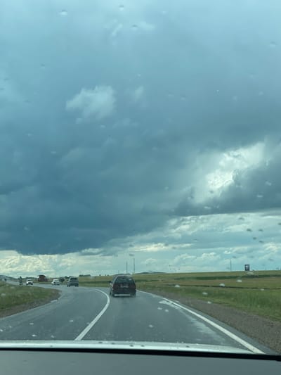 The image shows a view from inside a vehicle, looking out at a highway scene with a dark, cloudy sky. Water droplets are visible on the windshield. Several cars are driving on the road, which curves to the right. The landscape consists of green fields and distant trees, with some industrial structures and utility poles visible in the background. The sky is filled with dramatic, dark storm clouds, with patches of lighter sky peeking through. The overall impression is one of impending rain or a recent storm. The location is identified as being on the highway near the village of Zhañaaul, Kazakhstan.