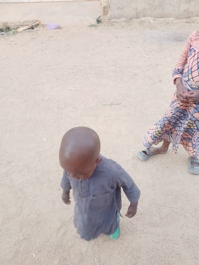 A child in a loose-fitting, gray garment stands on a tan, dusty ground. The child looks down, with a bald head visible. Part of another child wearing a patterned dress and gray shoes is to the right, standing still. The backdrop includes a building structure and scattered debris on the ground. The scene suggests an outdoor, possibly rural, environment.