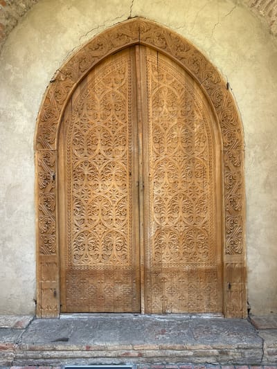 A closed, ornately carved, arched wooden door is set within an arched opening in a plastered wall. The door is comprised of two vertical halves. A pattern of carved circular and floral designs covers the majority of the door. Bands of geometrically repeating patterns border the design. Cracks run through the plaster above and around the door. To the right of the door, there is a brick column with a concrete coating. The scene is likely Sighnaghi, Georgia.