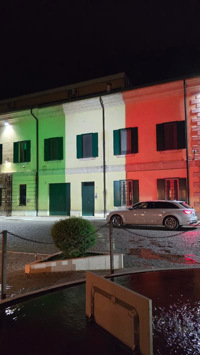 In the city of Peschiera del Garda, Italy, at night, a building is illuminated with the colors of the Italian flag: green, white, and red. The building features several windows with dark green shutters. A white Audi station wagon is parked on a cobblestone street in front of the building, which appears wet, suggesting recent rain. A small, round bush is planted in a raised planter box. In the foreground, a dark body of water, likely a fountain or canal, reflects the ambient light.
