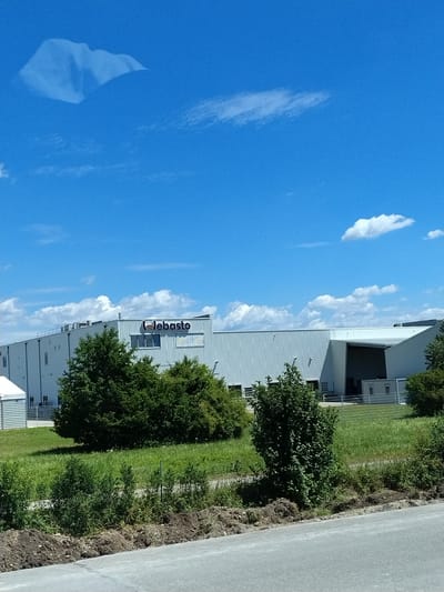 The image shows a large, modern industrial building with the "Webasto" logo visible on its facade. The building is white and appears to be constructed with corrugated metal siding. In front of the building, there is a patch of green grass with several trees. The sky is blue with scattered white clouds. The scene is likely an exterior shot of a Webasto facility in Hengersberg, Germany, based on the provided context. There are no people or specific actions depicted in the image. The environment suggests a sunny day.