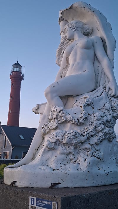 The image shows a white statue of a woman in the foreground, with a red lighthouse in the background. The statue is of a nude female figure, seated and partially covered by drapery and what appears to be a large shell. The lighthouse is tall and cylindrical, with a lantern room at the top. A building with a dark roof is visible behind the statue and to the left of the lighthouse. The sky is clear and blue. The statue is situated in Paldiski, Estonia, a town known for its historical significance and maritime connections. The signage on the base of the statue, partially visible at the bottom of the image, reads "LAEVA VIIMNE OHE LAST SIGH OF A SHIP", suggesting the statue may be related to maritime history or a shipwreck.