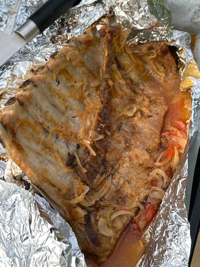 This close-up, high-angle shot captures a large portion of cooked meat, likely pork ribs, served within a crumpled aluminum foil container. The meat, showing clear bone structure, is golden-brown with some areas appearing slightly charred, indicating thorough cooking. Scattered over the meat are generous amounts of softened, translucent sliced onions and pieces of cooked tomato, which have melded into a glistening, savory liquid accumulated at the bottom of the foil. A knife with a black handle and a silver blade is partially visible in the bottom-left corner, suggesting the dish is ready for carving or serving. The aluminum foil, stained with cooking juices and fats, serves as a rustic baking or serving vessel. A dark, possibly black, edge of a baking tray or surface is visible along the top edge of the frame. The image focuses entirely on the food, conveying a sense of a recently prepared meal. There are no people visible, nor are there any explicit cues for time of day, weather, or emotions. No visible text is present in the image. The scene depicts a prepared meal, potentially from a domestic setting.