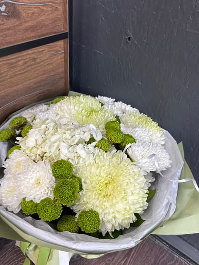 A bouquet of white and pale green chrysanthemums and small, round green flowers is displayed. The chrysanthemums have intricate, layered petals, with some displaying a yellowish hue towards their centers. The green flowers are densely packed with small florets, creating a textured, spherical shape. The bouquet is wrapped in pale green and white paper, with a delicate white ribbon tied around it.

The bouquet is placed on a surface that appears to be a wooden table or bench with a dark, textured background behind it. The wooden surface has a prominent dark stripe running down the middle. The background is a dark, slightly textured surface, possibly a wall or a countertop, with some subtle scuff marks visible.

The lighting suggests it is daytime, and the overall mood is serene and natural, highlighting the beauty of the flowers. There is no discernible text in the image. The setting does not provide strong cues about the specific location other than the wooden surface and dark background. The bouquet itself is the primary subject, arranged in a classic presentation.