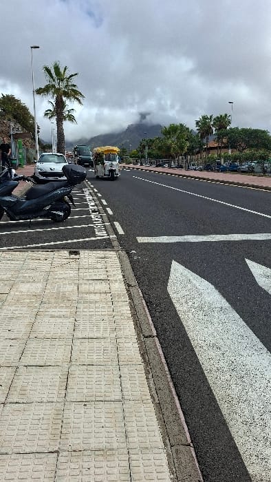 The scene depicts an urban street in Costa Adeje-playas Del Duque, Spain, under a heavily overcast sky with low, grey clouds obscuring the peaks of distant, dark mountains. In the foreground on the left, a light-colored, textured tiled sidewalk transitions to a dark grey curb. Adjacent to the curb, a paved shoulder marked with white dashed lines and partially visible white "BUS STOP" text contains a parked dark grey scooter with a black rear storage box.
The asphalt road to the right features white solid and dashed lane markings, along with prominent white directional arrows painted on its surface. Moving from left to right across the road, a white hatchback and a dark-colored SUV are positioned on the far left lane. A white three-wheeled auto-rickshaw with a yellow canopy is observed traveling rightward in the primary lane of traffic. On the far left, partially obscured by a street light and green foliage, a person in dark clothing is visible. The right side of the road is bordered by a wide paved area containing numerous parked scooters and motorcycles, backed by a line of tall palm trees and indistinct low-rise buildings. Multiple streetlights are visible along both sides of the road.