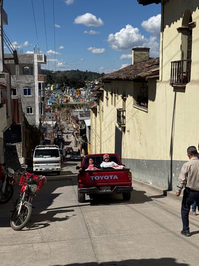 A sunny, tilted street scene features a red Toyota pickup truck with three people riding in the bed. A white truck follows. A red motorcycle with a basket is parked near the curb. Buildings of various heights line the narrow street, leading to a distant town nestled in the hills under a blue sky with white clouds. A person is walking along the sidewalk at the top of the frame.