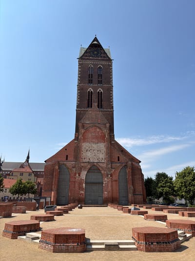 This image depicts the St. Nikolai Church in Wismar, Germany. The red brick church has a tall, imposing tower with a clock face and a pitched roof. The church's facade features large, arched windows and weathered brickwork. In front of the church, there is an outdoor plaza with circular, stepped brick structures and a spherical object. The sky is clear and blue, with a few wispy clouds. Trees and other buildings are visible in the background.