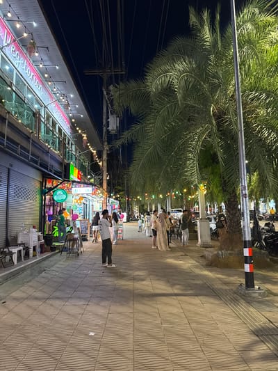 The image depicts a street scene at night in Ban Klong Haeng, Thailand, likely a commercial area with restaurants and shops. The scene is illuminated by streetlights, string lights, and the lights from the establishments, creating a lively atmosphere.

The main subjects are people walking and standing on the sidewalk, which is paved with wavy patterned tiles. Some individuals are dressed in casual attire, while others appear to be tourists, possibly in traditional or ceremonial clothing, suggesting an event or celebration. There are also motorbikes parked along the street.

Notable details include large palm trees lining the sidewalk, adding a tropical feel to the setting. Colorful string lights are strung across the street and above the storefronts, enhancing the festive ambiance. Signs for businesses are visible, with one prominent banner displaying the text "Vegetarian Restaurant" in stylized pink lettering. Other signs include prices in Thai baht, such as "High 100 B" and "50 B". The time of day is clearly night, with a dark sky and artificial lighting dominating the scene. The weather appears to be clear, allowing for outdoor activity.

The overall mood of the image is vibrant and social, suggesting a bustling evening in a popular tourist or local gathering spot. The presence of people of different ages and attire hints at a diverse crowd enjoying the evening.