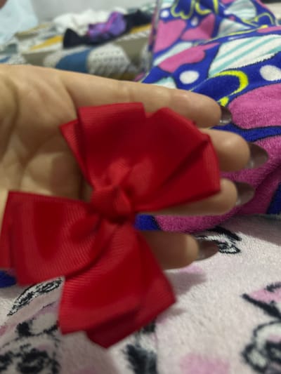 A red ribbon bow is held in a person's left hand. The fingernails are long and painted a dark color. The background includes colorful patterned fabrics. The location is unknown.