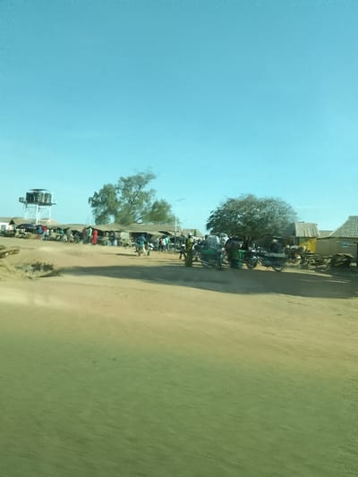 The image depicts an unpaved market area in Mangu Halle, Nigeria, under a clear, bright blue sky. The immediate foreground displays a blurred green and brown surface, indicative of a view from a moving vehicle over a dirt road. In the mid-ground, a bustling market is visible, comprising multiple low-profile, rustic vendor stalls with corrugated metal roofs. Numerous individuals are interspersed throughout this market space, engaged in various activities. On the far left, a tall, open-frame metallic structure, possibly a water tower or communication tower, rises above the surrounding buildings. Several motorcycles are present on the unpaved ground, both parked and in motion, with at least one motorized tricycle (keke Napep) also visible on the right side. Scattered trees, including one with sparse foliage on the left and a more densely leafed tree on the right, define the background against a backdrop of additional modest structures with corrugated roofs.