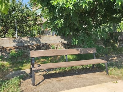 A wooden park bench with black metal legs sits on a dirt path in front of a low stone wall. Behind the wall, a chain-link fence encloses trees and foliage. A building with green and white awnings is visible through the trees. The scene is outdoors in daylight.