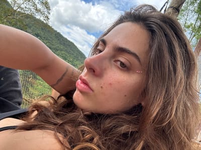 A woman's face dominates the frame, her head tilted. She has light brown hair and fair skin with three pink jewels near her eye. Her lips are full and pink. A faint tattoo is visible on her arm. The background is a mix of nature and structures; the sky is visible with white clouds, a lush green mountain slopes up, and there is part of what appears to be a fenced area.