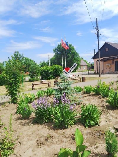 The image depicts a sunlit scene in the city of Изыхские Копи, Russia, featuring a decorative signpost, flags, and landscaping. The signpost, shaped like a diagonal bar with a red heart at the beginning, reads "КОПИ ИЗЫХСКИЕ" in white lettering, indicating the location. Two flags, one red and one with the Russian tricolor, are planted behind the signpost.
The environment is a public outdoor space, possibly a park or a town square. In the foreground, there is a well-maintained flower bed with lush green plants and clusters of purple flowers, interspersed with a small evergreen tree. A pathway, paved with light-colored material, runs through the scene.
Behind the flower bed, a brown wooden bench with decorative metal supports is visible. Further in the background, there are more trees, including pine trees and dense green bushes. A light-colored building with a brown roof and windows is situated on the right side of the image, likely a public facility such as a post office or administrative building, with a notice board on its exterior. A utility pole with electrical wires stands near the building. Residential houses with green roofs can be seen in the distance. The sky is blue with scattered white clouds, suggesting fair weather. No people are visible in the image.