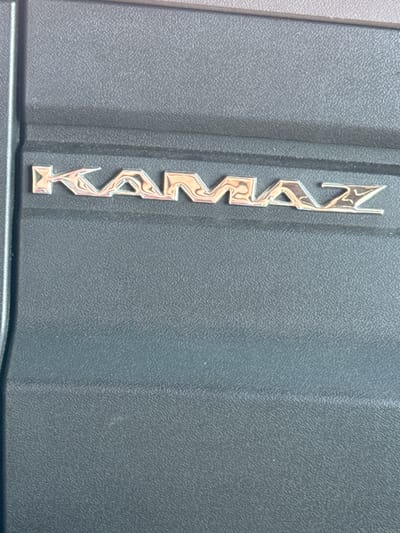 The image shows a close-up, vertical view of a silver metallic emblem with the word "KAMAZ" embossed on a textured, dark gray surface. The emblem is comprised of individual, stylized letters that appear to be chrome or polished metal, reflecting light and creating a shiny, three-dimensional effect. The "K" is at the top, followed by "A", "M", "A", "Z", and finally what appears to be a stylized "Z" or a symbol below it. The background surface has a fine, pebbled texture, characteristic of plastic or a similar material often used in vehicle interiors or exteriors. A subtle vertical seam runs down the right side of the frame, dividing the textured surface into slightly different planes. The lighting suggests an outdoor setting, likely daylight, given the bright reflections on the metal. The image is cropped tightly on the emblem and the immediate surrounding surface, providing no wider context of the environment or any people. The focus is solely on the branding of the vehicle. There is no discernible time of day, weather, or emotional cues. The location context is Sim, Russia, but this information is not visually evident from the image itself.