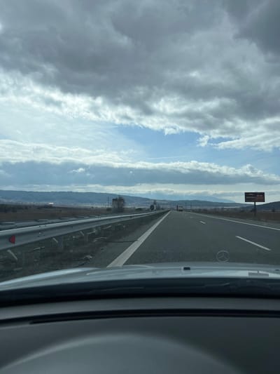 The image presents a forward-facing, uncropped view from inside a vehicle traveling on a multi-lane highway, near the city of Vladimir, Bulgaria. The immediate foreground shows a portion of the vehicle's dark-toned dashboard and the silver hood, featuring a prominent Mercedes-Benz emblem.

The highway stretches into the distance, appearing as a smooth asphalt surface with clear lane markings, including a solid white line separating the left lane from the shoulder, and a dashed white line further right. To the left of the highway, a metal guardrail with red circular reflectors runs parallel to the road. Beyond the guardrail, the terrain consists of dry, sparse groundcover.

In the midground on the right side of the highway, a large rectangular billboard is visible, displaying Cyrillic text and an advertisement, possibly for food or a restaurant, featuring an image with red hues. Another metal guardrail is present further along the right side of the road.

The background is dominated by a long, undulating range of hills or low mountains, partially covered with sparse, possibly deciduous, vegetation or bare trees, under a mixed sky. The sky above is characterized by significant grey cloud cover, interspersed with distinct patches of clear blue sky, particularly in the mid-upper section. Two faint, linear contrails are visible within the blue portions of the sky. Several large commercial trucks are discernible in the far distance on the highway, traveling in the same direction as the vantage point. No individuals are visible in the scene.