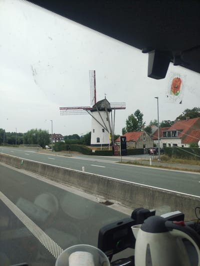 An exterior, eye-level shot captures a classic white windmill with red-trimmed sails, located in Hoeke, Belgium. The windmill is situated amidst a cluster of buildings with red-tiled roofs, surrounded by green trees. A gray asphalt road with white lane markings runs in the foreground, separated by a concrete barrier. Reflections from the interior of a vehicle are visible on the windshield, including a black device and a white electric kettle. The sky is overcast and appears bright white.