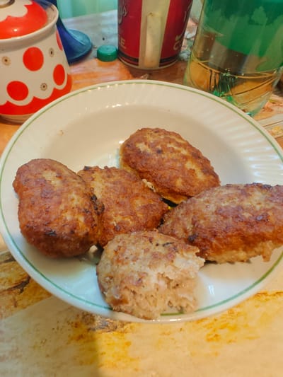 A close-up, overhead shot shows a white, shallow bowl with a green rim filled with five golden-brown, pan-fried cutlets, arranged in a cluster. One cutlet in the foreground has a bite taken out of it, revealing a pale, textured interior. The bowl rests on a light-colored, patterned surface. In the background, to the left, is a white ceramic container with red polka dots and a scalloped red rim, possibly a sugar bowl or canister. Next to it, a dark blue object, possibly a funnel or lid, is visible. Behind the cutlets, to the right, is a clear plastic bottle filled with a pale yellow liquid, with green text on the label. Further back, partially visible, is a red and white cylindrical object, likely a can. The overall setting appears to be a home kitchen or dining area. The location is Pryvokzalne, Ukraine.