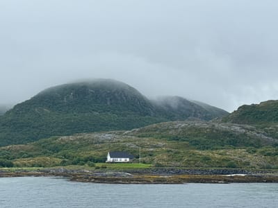 The image depicts a cloudy, overcast day in Onøya, Norway, with a body of water in the foreground. Beyond the water, a rocky shoreline leads to a lush green hillside. Further inland, a white house with a dark roof is situated on a grassy knoll. Behind the house, rolling hills covered in dense green vegetation rise up, disappearing into the fog-shrouded mountains in the background. The overall atmosphere is serene and somewhat desolate.