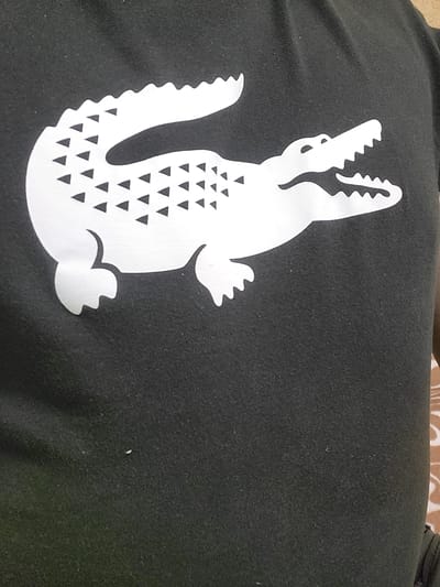 A close-up shot of a dark fabric, possibly a t-shirt, displays a large white graphic of a crocodile. The crocodile is positioned in the center of the frame, facing to the right. Its body is outlined in white, and its scales are depicted by numerous small, black triangles arranged in rows along its back and tail. The crocodile's mouth is open, revealing jagged white teeth. The fabric itself appears to be textured, with a slight sheen that suggests it might be a synthetic blend. The background is entirely filled with the dark fabric, with no other discernible elements or environmental context. The image does not provide any information about the location in Khotsky, Ukraine.