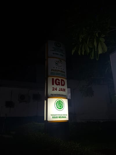 The image shows a vertical illuminated sign for RSUD Meuraxa in Banda Aceh, Indonesia. The sign is positioned at night in what appears to be an outdoor setting, with a dark background and some foliage visible at the top right. The sign itself is segmented into different sections. The top section displays "RSUD MEURAXA BANDA ACEH" followed by an address and a phone number. Below this is a section indicating "PARIPURNA" with stars and text about accreditation. The largest section in the middle prominently displays "IGD 24 JAM" (Emergency Room 24 Hours). The bottom section shows a logo with "MAJELIS ULAMA INDONESIA" and text indicating "TERSITIFIKASI DSN-MUI RSUD MEURAXA". The overall scene is dark, with the illuminated sign being the primary source of light, highlighting the information on it. There are no people visible in the image. The location is identified as being in the city of Lampeuneurutblang, Indonesia based on the metadata.