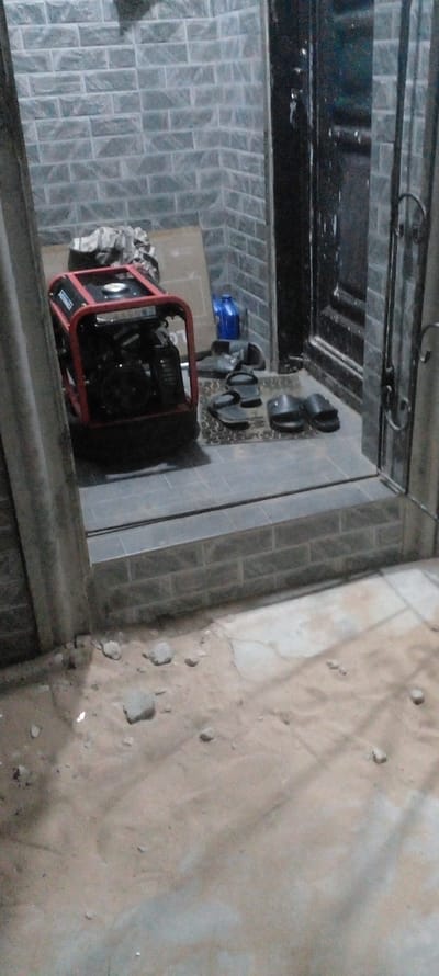 The scene, seemingly taken from an elevated position in Agunmo, Nigeria, depicts an outdoor utility area adjacent to a building. The ground is partially covered in a sandy, light brown surface with scattered rocks or debris. Along the edge of the building, a tiled walkway with a gray brick pattern is visible. Near a doorway, a red-framed generator sits on a light-colored, possibly wooden, platform. Next to the generator are various objects, including what appear to be black, possibly plastic, pipe fittings and a blue container. The doorway itself is dark, with a metallic frame. The overall impression is of a utility space, possibly for storing equipment and carrying out maintenance, located in an urban or peri-urban environment. No people are visible.