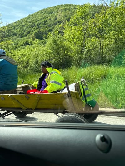 A view from inside a vehicle shows a scene of workers beside a road. Two individuals are visible, one wearing a high-visibility yellow jacket and black helmet, appearing to be working inside a yellow cart or trailer. There is another individual wearing a blue shirt standing adjacent to the cart. The cart has wheels, and atop it rests another high-visibility yellow vest and other objects. The background includes green foliage and a partially visible hillside covered with trees. The image is taken at an angle. Given the information provided and the scenery, the location could be in or near Bilka, Bulgaria.