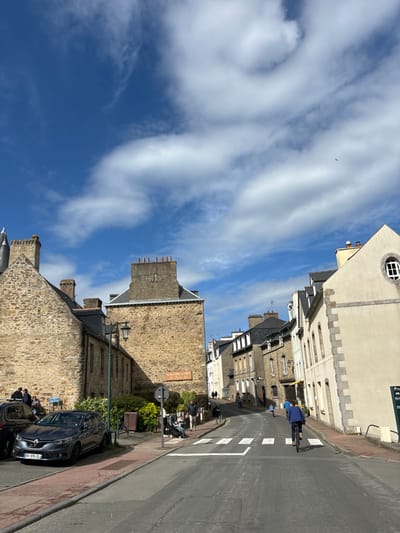 The image shows a street in Dinard, France, specifically at the coordinates 48.631, -2.053. The scene is a narrow street with stone buildings on both sides. The sky is a bright blue with fluffy white clouds. The street is paved and has white painted crosswalks. There are several people walking on the sidewalk, and a man is riding a bicycle down the center of the street. There are a couple of cars parked on the side of the street.  The stone buildings are old and have a variety of architectural details, including windows, doors, and chimneys. The buildings are mostly in good condition, but there are some signs of wear and tear. There are several trees and shrubs along the sides of the street, adding some greenery to the scene.