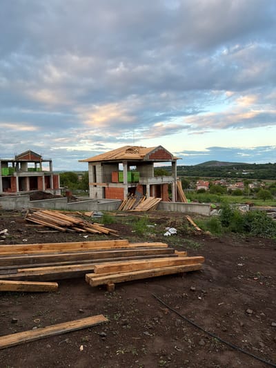 The image shows a construction site in Cherni Vrah, Bulgaria. Partially constructed buildings with red brick walls and concrete supports are visible. Wood planks are stacked on the ground beside the buildings. The sky is cloudy, and there are houses and hills in the background. No people are present. A black hose lies on the dirt.