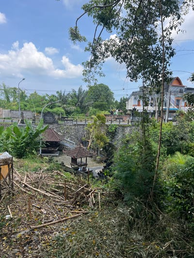 The image depicts a landscape in Buduk, Indonesia, featuring buildings, vegetation, and architectural structures. The sky is visible with scattered clouds. In the middle ground, there are buildings, and further down, structures with tiered levels are visible, constructed with stone and traditional roofing. To the right, there is a pile of dried bamboo. Vegetation is dense throughout the scene. There is an electrical pole on the bottom left.