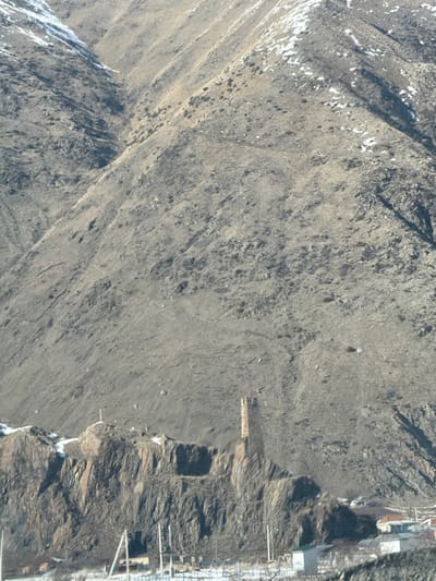 This image presents a wide view of a mountainous landscape in Achkhoti, Georgia, under clear daylight conditions, suggesting a cold season due to visible snow. The upper two-thirds of the frame are occupied by a steep, rugged mountain face, predominantly grey and brown, with minimal vegetation and scattered white patches of snow in depressions and higher areas. Evidence of erosion is visible across the slope.
In the lower third of the image, a tall, weathered stone tower with a rectangular base stands prominently on a rocky outcrop, which also features dark, fractured rock faces. To the left of the tower, on a lower peak of the same outcrop, a white cross is visible. Below the rocky formation and extending into the foreground, a small rural settlement is discernible, consisting of multiple buildings with varying roof colors, interspersed with patches of snow on the ground. Modern infrastructure, including utility poles and overhead power lines, crosses the immediate foreground. No individuals or specific actions are visible within the frame.