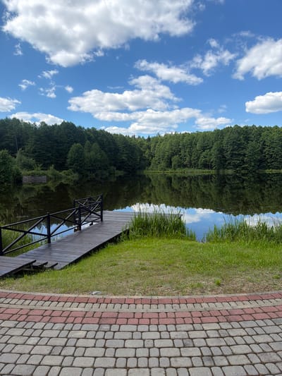 The image depicts a serene lakeside scene at a location within the Ляснянскі сельскі Савет, Belarus. In the foreground, a paved walkway with a pattern of gray and red interlocking bricks curves towards the right. Beyond the walkway, a patch of vibrant green grass leads to a wooden pier extending over the dark, still water of a lake. The pier is constructed of dark wooden planks and features a black metal railing with a decorative crisscross pattern. Tall, green reeds grow along the edge of the lake near the pier. The lake reflects the lush green forest that lines its opposite bank, creating a mirror image of the trees and the sky above. The sky is a clear blue, punctuated by fluffy white cumulus clouds. The overall impression is one of tranquility and natural beauty. There are no people visible in the image.