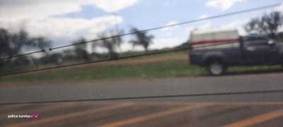 A blurred, wide-angle shot shows a roadside scene with a focus on foreground elements. Utility lines stretch across the frame, partially obscuring the view. A pickup truck with a camper shell is parked on the right side of the road. Beyond the road, a field of green grass leads to distant trees under a cloudy sky. The overall image quality is poor due to the blurriness. "police komba" and a heart emoji are added in the bottom left corner.