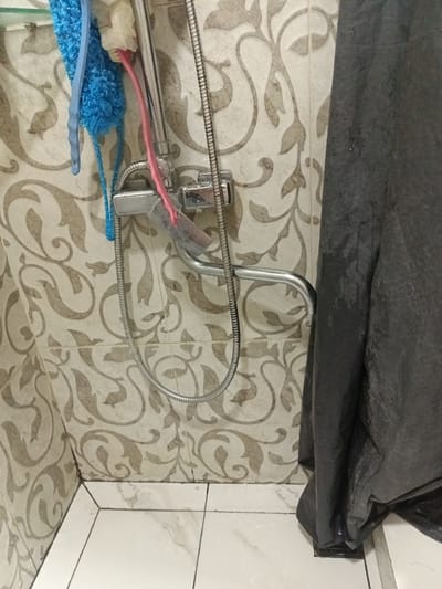 In a bathroom in Yukariyuz, Uzbekistan, a close-up shot reveals a shower area. A chrome shower fixture with a flexible hose is mounted on a tiled wall. Hanging from the fixture are several bath accessories, including a blue loofah and a pink-handled brush with a white sponge. To the right, a dark grey shower curtain is partially drawn, obscuring the rest of the shower stall. The tiled walls have a patterned design in beige and brown. The floor is tiled with white marble-patterned tiles. Water droplets are visible on the tiles and the shower curtain, suggesting recent use.