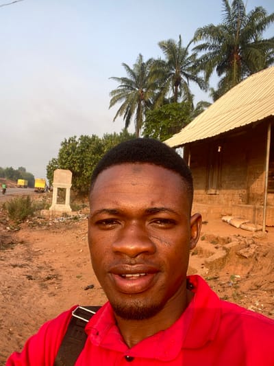 A man wearing a red shirt smiles into the camera. He is outside on a dirt road next to trees and some buildings. The sky is light blue and there are trucks in the distance. The photo is slightly blurry and the man's skin is shiny.
