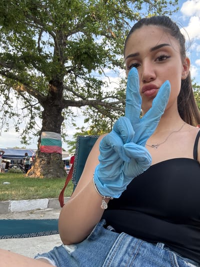 A young woman is prominently featured in the foreground of an outdoor scene in Kostievo, Bulgaria. She is seated, facing the viewer, with her head slightly tilted and her lips pursed in a pout. Her left hand, encased in a light blue disposable glove, is raised, forming a "peace sign" gesture. She has long, dark hair pulled back, and her facial features include dark eyebrows and visible makeup. She wears a black spaghetti-strap top, denim shorts, a silver necklace with two small heart pendants, and a silver charm bracelet on her left wrist. She is seated in a dark green folding chair with a geometric pattern.
In the background, a large, mature tree with dense green foliage occupies the left side of the frame. Wrapped around the lower trunk of this tree is a fabric band displaying the horizontal stripes of white, green, and red, consistent with the flag of Bulgaria. To the left and behind the tree, an outdoor gathering is visible, featuring multiple individuals under blue and white canopies or tents. These individuals are engaged in various activities, some standing and others seated. The ground consists of green grass and a light-colored paved curb. The sky is bright blue with scattered white clouds, indicating daytime conditions.