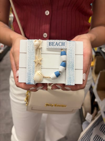 A person is holding a decorative sign that says "BEACH". The sign is made of white wood planks and decorated with shells, a starfish, and blue and white buoys. The person is wearing a maroon shirt and white pants. They are also carrying a small beige purse with gold lettering. The background is blurred, but it appears to be an indoor setting, possibly a store. There is no visible indication that this scene is located in Würzburg, Germany.