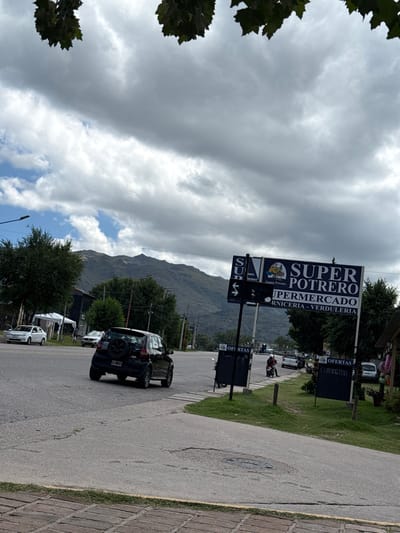 A street scene under a cloudy sky shows a "SUPER POTRERO" supermarket sign with directional arrows. Cars are parked and driving along the road. Mountains are visible in the background. Sidewalk is made of bricks. Additional offer signs can be seen near the supermarket advertising. The trees line the road.