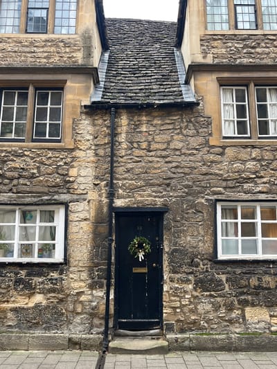 The image displays the stone facade of a building located in Oxford, United Kingdom. The structure is characterized by walls constructed from irregular, light-brown and grey stone blocks. A steep, dark grey slate roof, showing signs of moss or lichen growth, is visible in the upper-center, connecting two sections of the building.
Four windows are present: two upper-level, multi-pane windows with dark frames, positioned above the main wall sections to the left and right. Below these, two white-framed, multi-pane casement windows flank a central entrance. The lower-left window contains internal decorations, including a circular peace symbol and a smaller, square stained-glass-like ornament. The lower-right window is obscured by white curtains.
A black vertical downpipe runs along the stone wall, positioned to the left of the doorway, extending from beneath the roofline to the ground. The central entrance features a dark, likely black, wooden door with the number "13" affixed above it. A brass letterbox and a dark handle are visible on the door. A circular wreath, composed of green foliage, pinecones, gold and white spherical ornaments, and a white ribbon, hangs centrally on the door.
The building rests on a grey paved surface, with a narrow metal grate or drain cover visible near the base of the wall. The sky above is overcast. No individuals are present, and no actions or interactions are depicted.
